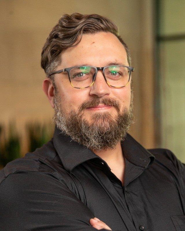 Adam Valine, Co-Founder of HelloDave.ai, turned slightly towards the camera with his arms folded. He has swept-back brown hair, a full brown beard and tortoiseshell glasses, and wears a black button-down shirt.