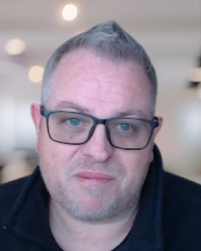 Dean Brown, Founder of HelloDave.ai, facing the camera in a bright open-plan office. He has short grey hair, a trimmed grey beard and dark rectangular glasses, and wears a dark zip-neck top.